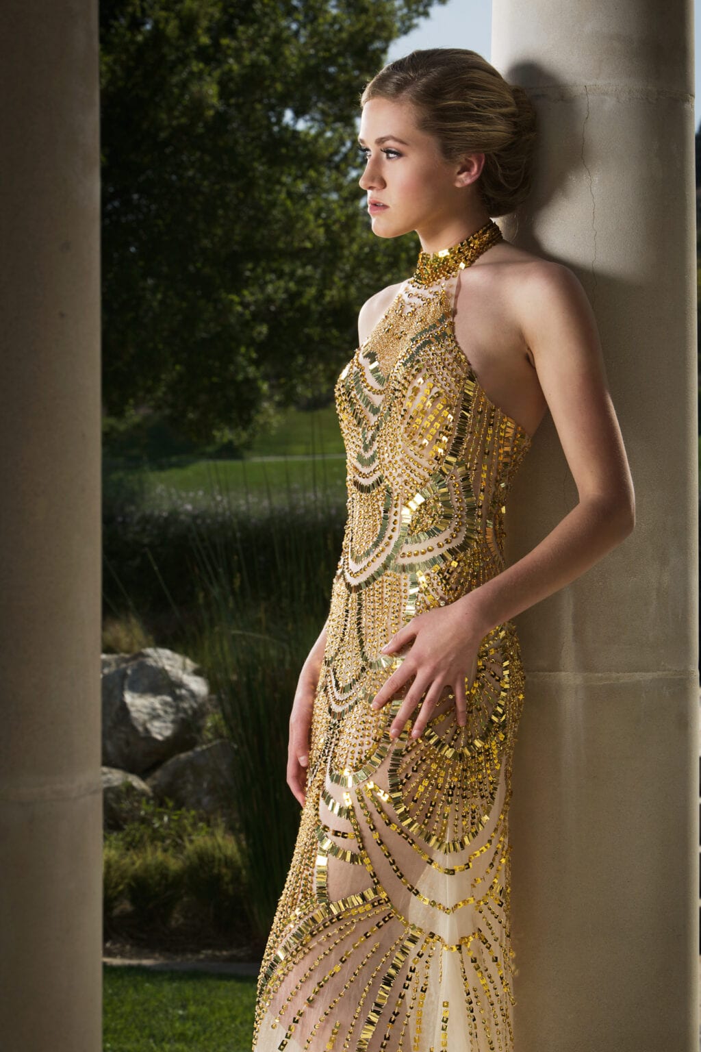 Model in a gold beaded halter gown leaning against a column.
