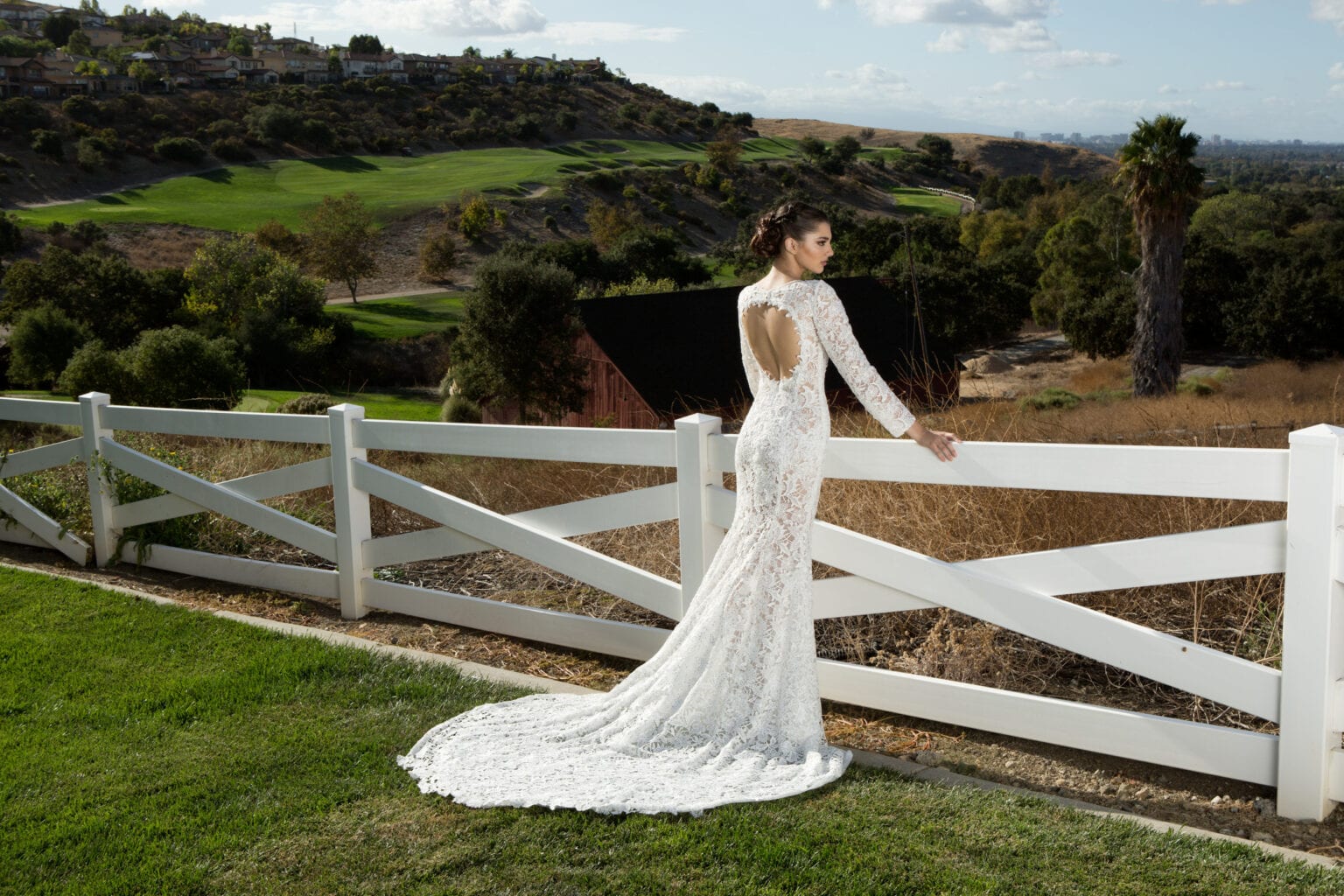 Model in a lace bridal gown with a long train beside a white fence.