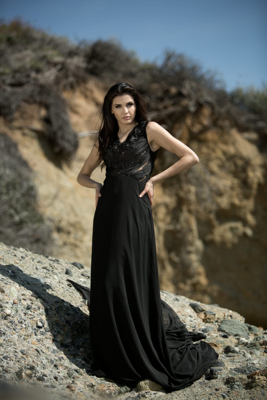 Model in a long black gown standing on rocky terrain.
